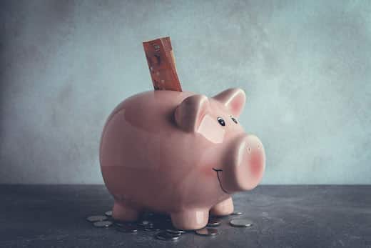 Benefits betriebliche Altersvorsorge | Rosa Sparschwein auf grauem Tisch mit grauem Hintergrund mit Geldschein und Münzen drum herum
