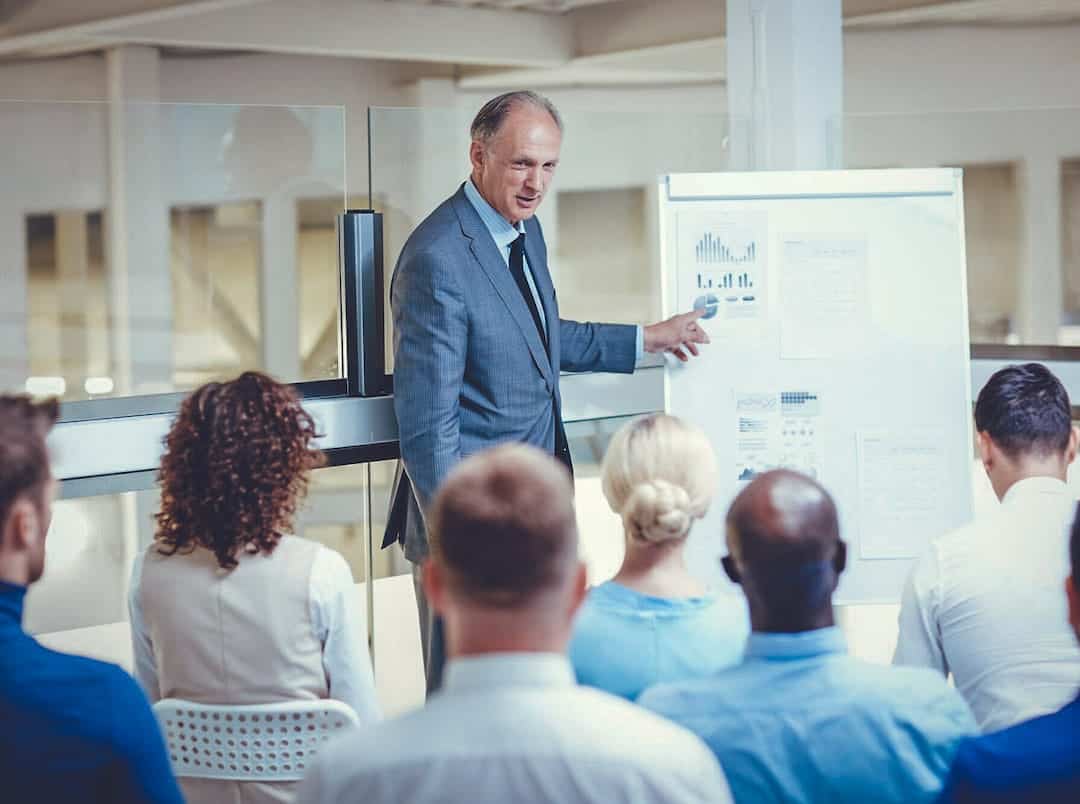 Mehrwerte von Benefits für Arbeitgeber und Arbeitnehmer | Mann im Anzug steht vor einer Gruppe sitzender Menschen mit einer Tafel mit Diagrammen